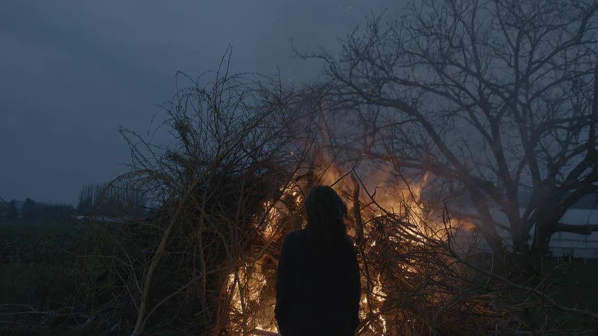 A person stands in front of a fire burning a big pile of branches