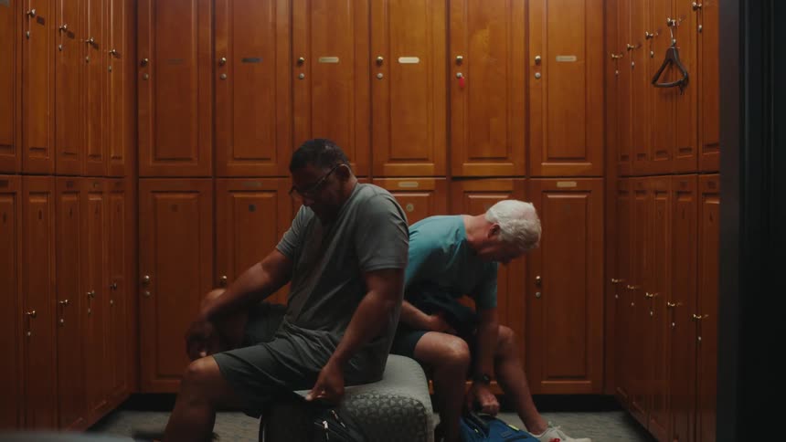 Two men in a locker room picking up bags and putting on equipment