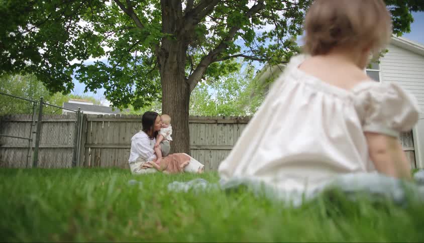 Two small children sitting on the grass in a backyard and in the background the mother holding a baby next to a tree