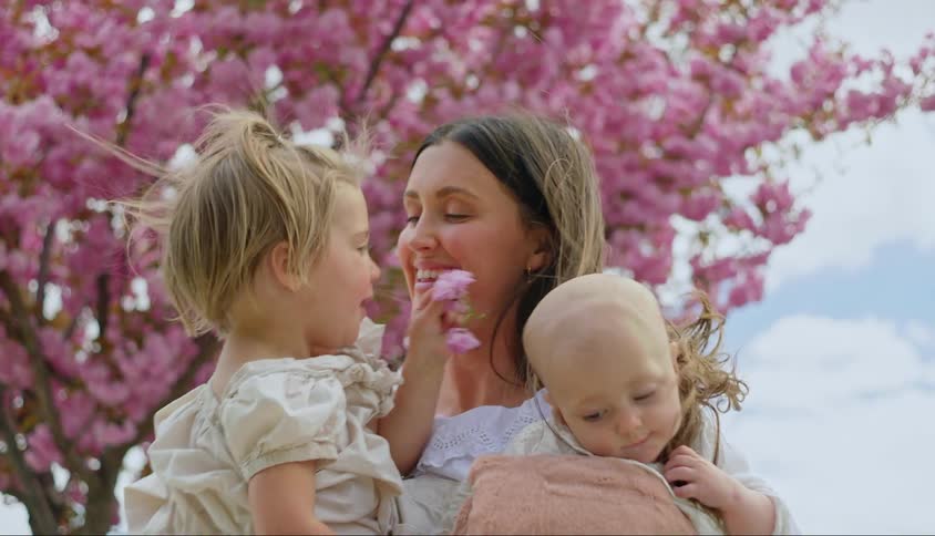 A mother standing in front of a cherry blossom tree trying to hold both her young children