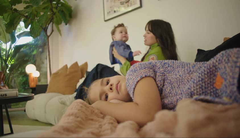 A mother bounces her infant son on a couch as her daughter lies on a blanket looking bored