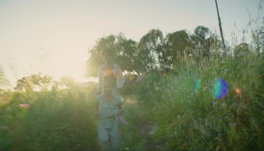 A mother and daughter laugh while running through a grassy area choked with weeds