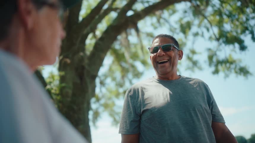 Man laughs while talking with a friend as they stand near a large tree