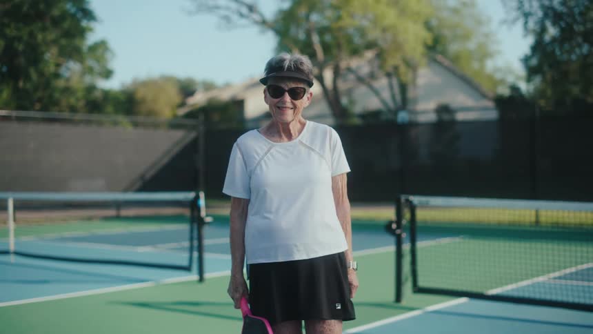 Elderly lady with sunglasses holding a pickleball paddle on a tennis court