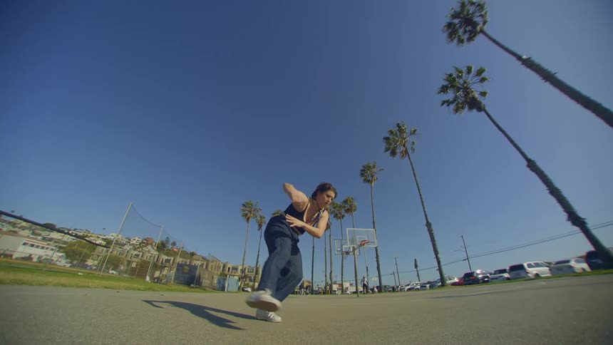 A woman doing dance moves on a concrete floor on a clear day with palm trees on the right side