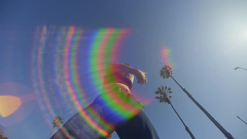 A woman doing dance moves with her arms and there is a flare of colored light in front of her coming from the blue sky