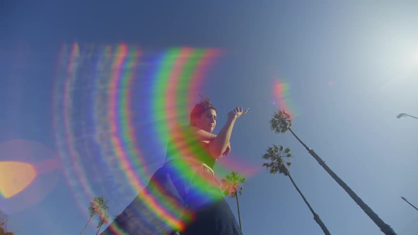 A woman standing making gestures with her arms in contrast to the blue sky and a colorful flare in front of her