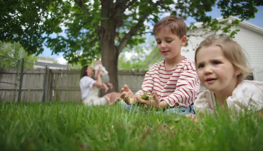 Two kids goof off while their mother bounces a baby near a tree in a yard