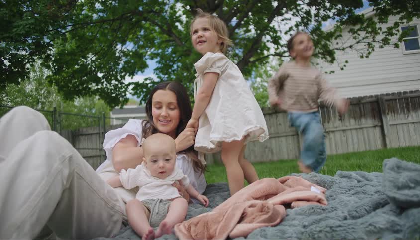 Mother lying down with baby and a toddler and next to a little boy running while they are on the grass in the backyard of a house