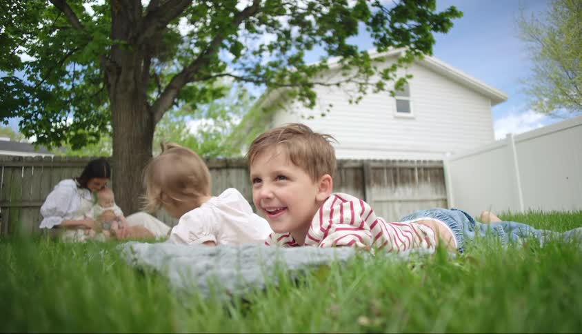 Two children lay on blanket in grass mother holds baby under tree