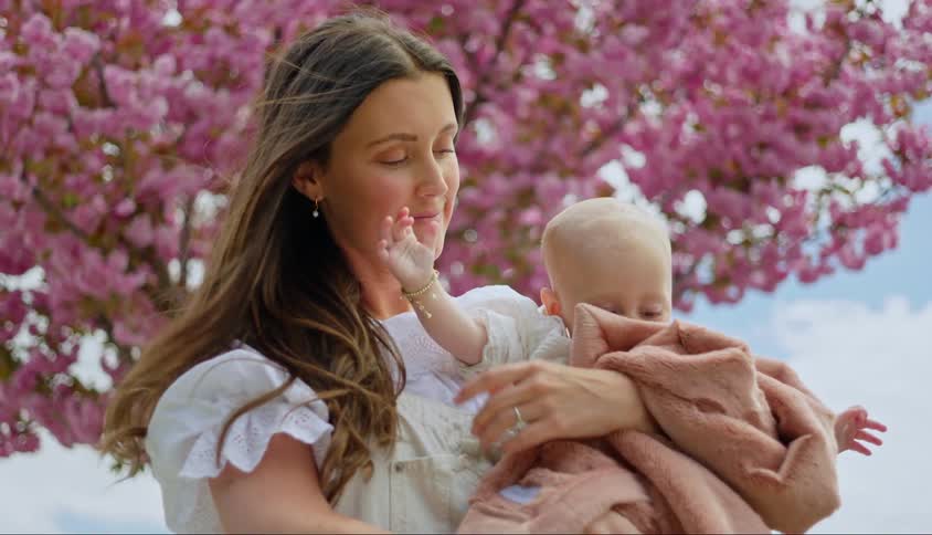 Woman standing outside near a flowering tree on a windy day smiles and kisses her baby
