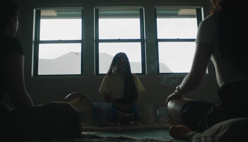 Three individuals in a dimly lit room with two windows perform rituals of meditation