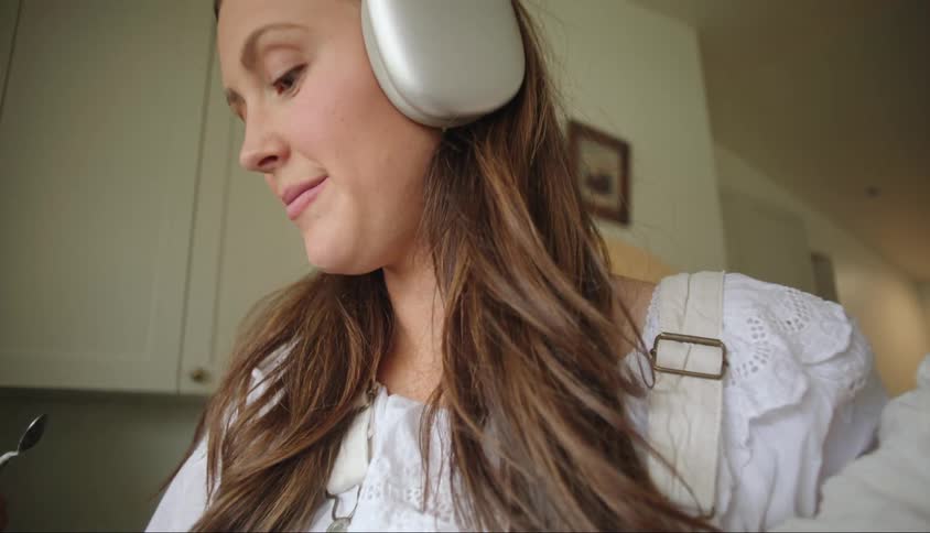 A mother talks to a child while holding a baby and listening to headphones in the kitchen