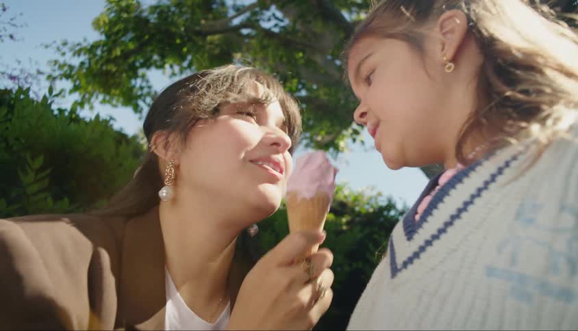 Mother crouches to share ice cream cone with her daughter while outdoors near a tree