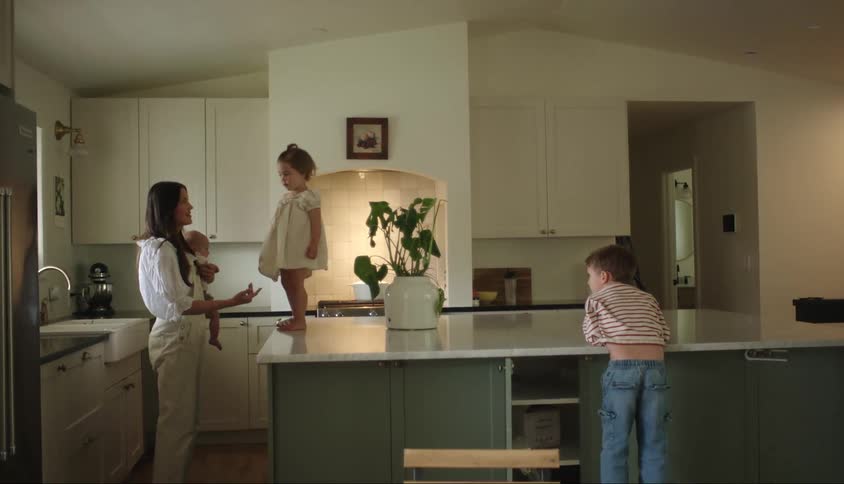 Mother in the kitchen holding a little girl on her lap holding a baby and her other son playing next to her