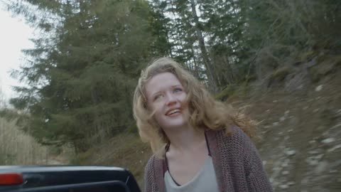A woman rides in the back of an open bed truck going through a forest