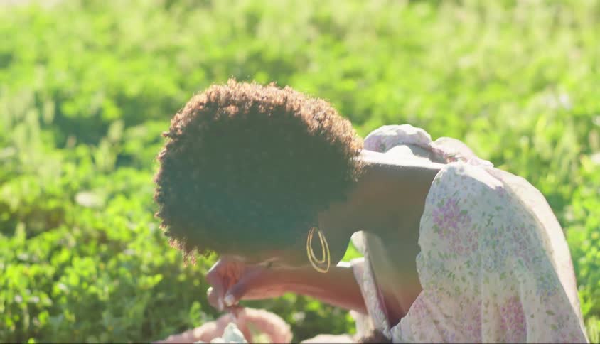 A mother tickles her young daughter with a plant stem while they are on a blanket in a grassy field