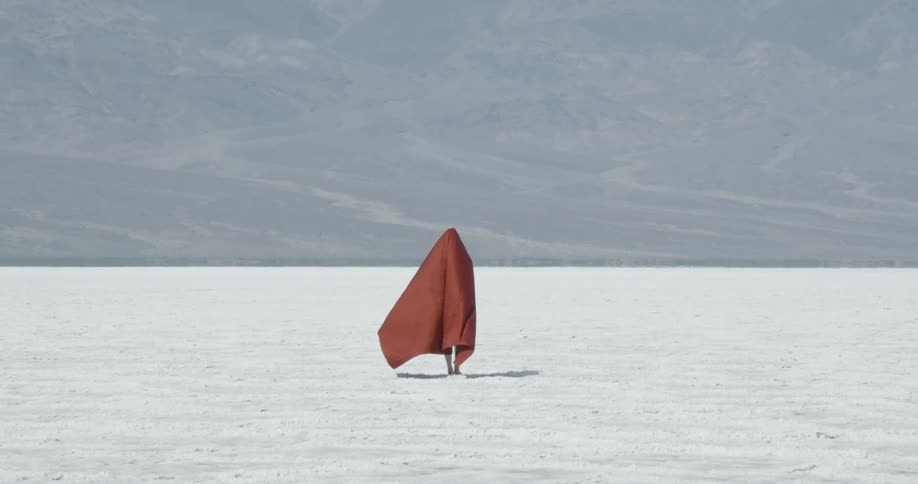 A person wearing a red cape over their head walks in the white sandy desert