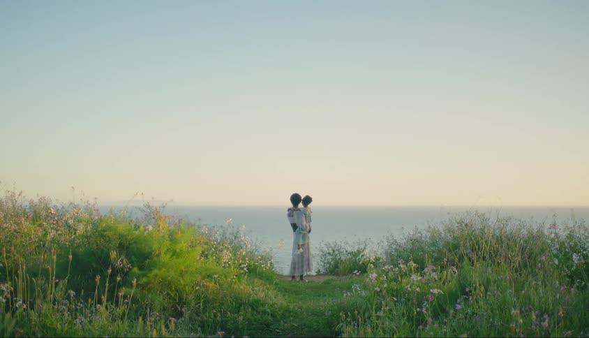 A mother and daughter looking out at the ocean from a grassy field on a coast