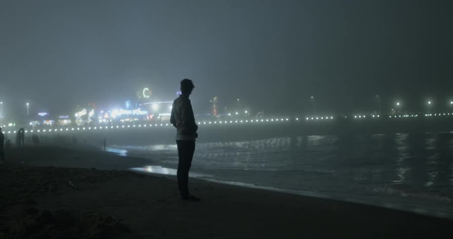 Man standing at the shoreline with an active pier in the background at night