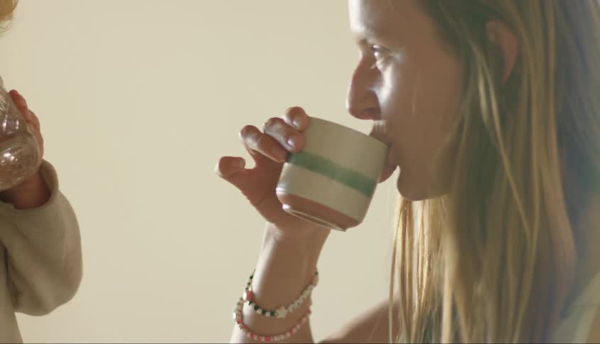 A blonde woman drinking from a mug and looking at a little boy in front of her drinking water from a glass cup