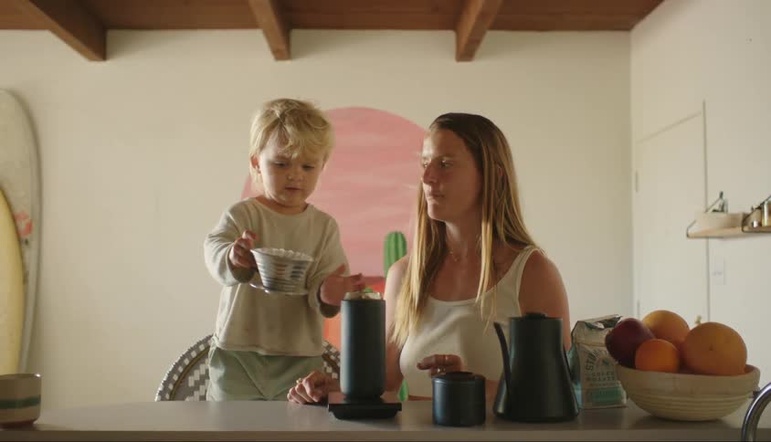 A mother pulling out a black bottle of coffee and a little blond boy taking out an object that is on top of the bottle