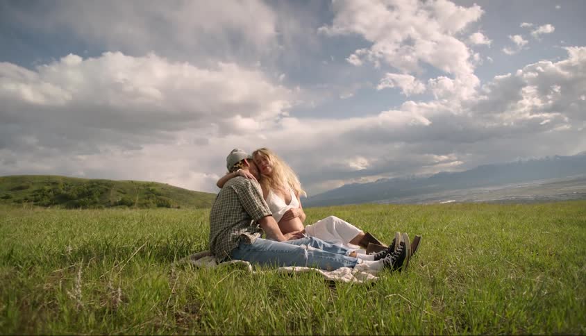 Man and woman hugging while sitting in a grassy field under a partly cloudy sky