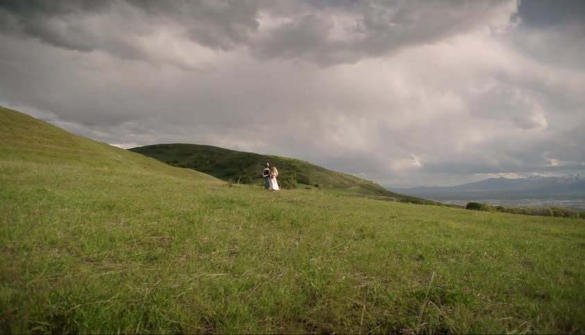 A couple is holding hands running in a field of green grass with hills in the background