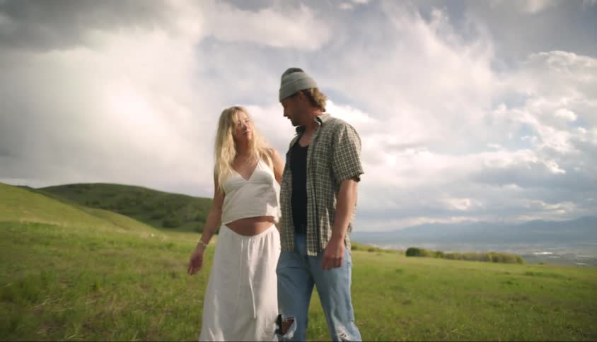 A man holds hands with a pregnant woman and they are both walking in a grass field