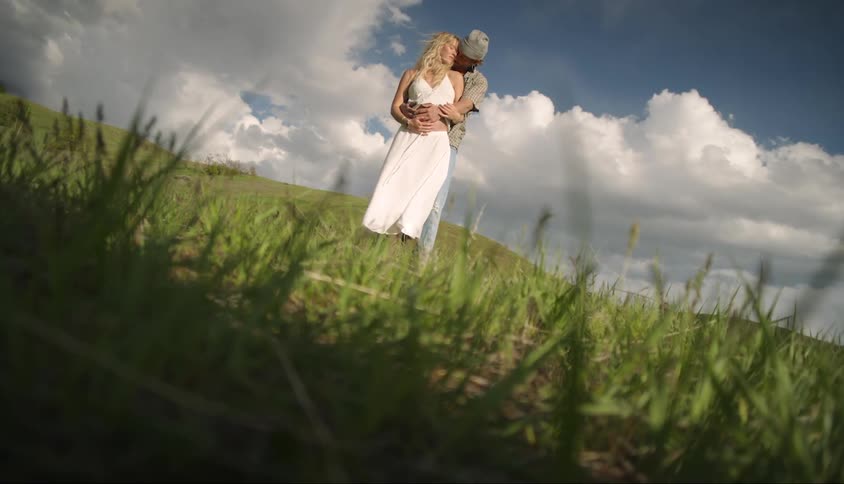 Man hugging a pregnant woman wearing a white dress in a field