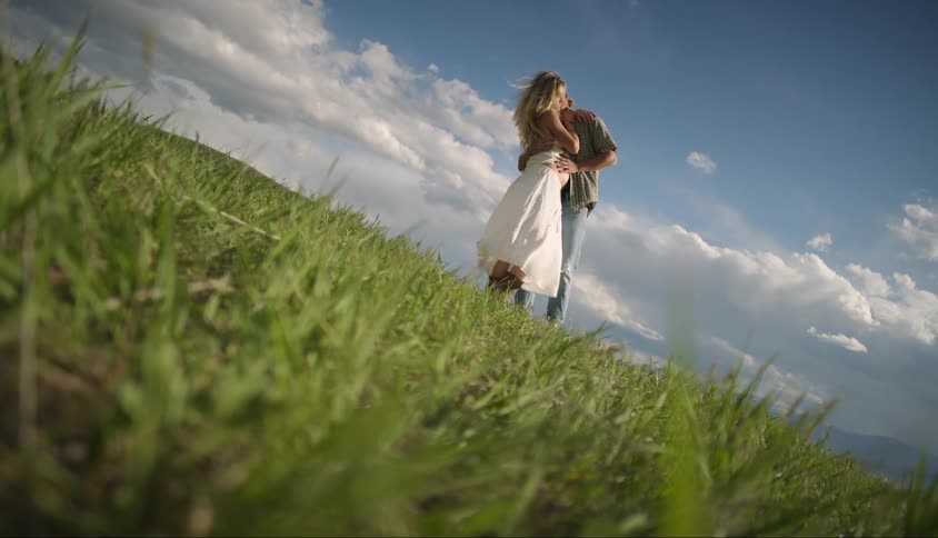 A man hugs and kisses a pregnant woman in a grassy field