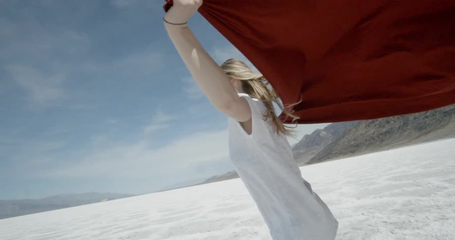 Woman running in white sand with a large red cloth flowing from her hands that are spread out to her sides