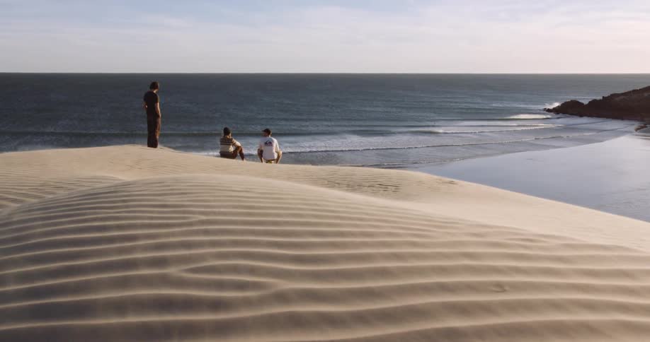 Three people stand on a sand hill watching the sea on an empty beach
