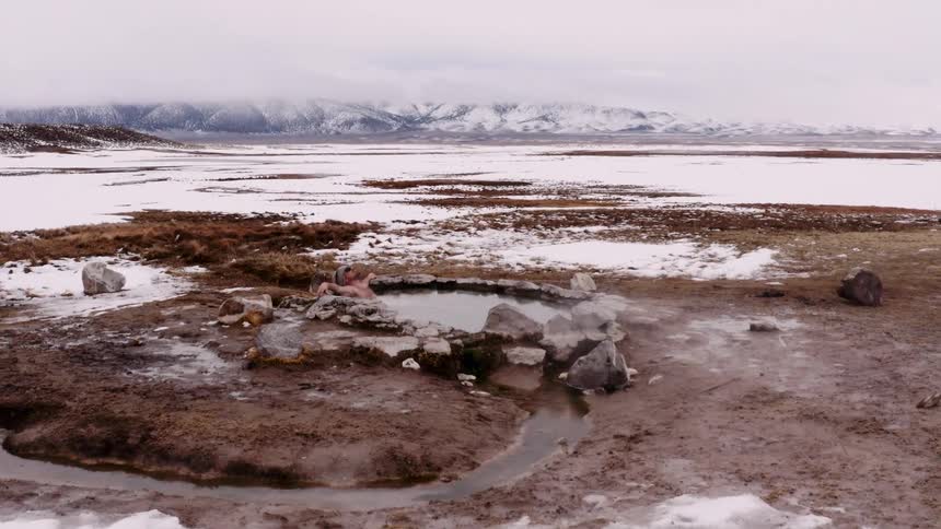 Person at a geothermal spring in a vast open landscape with patches of snow and distant mountains