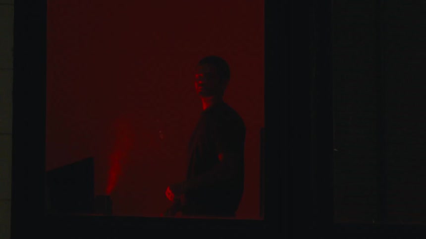 Man at the window of an apartment in a dark room with red light closing the curtain