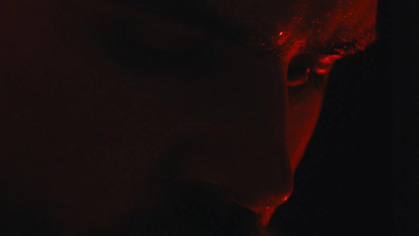 Closeup of a man in the dark one side ofhis face is illuminated as he looks down and a drop of sweat falls from his nose as he blinks