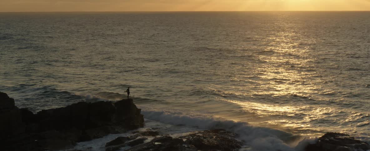 A surfer stand in silhouette on a coastal rocky outcropping