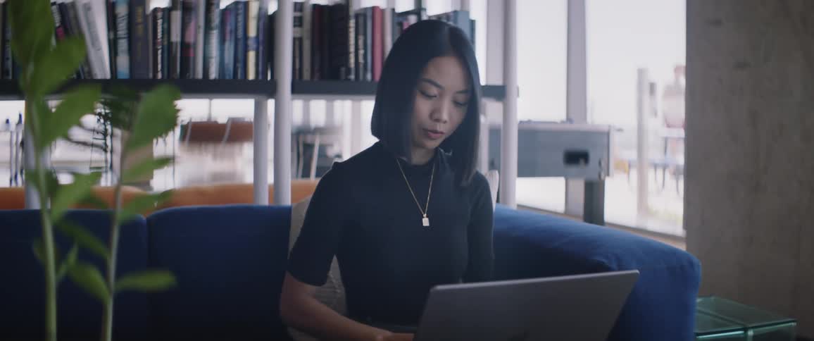 Woman sitting near shelved books smiles and talks while she types on a laptop