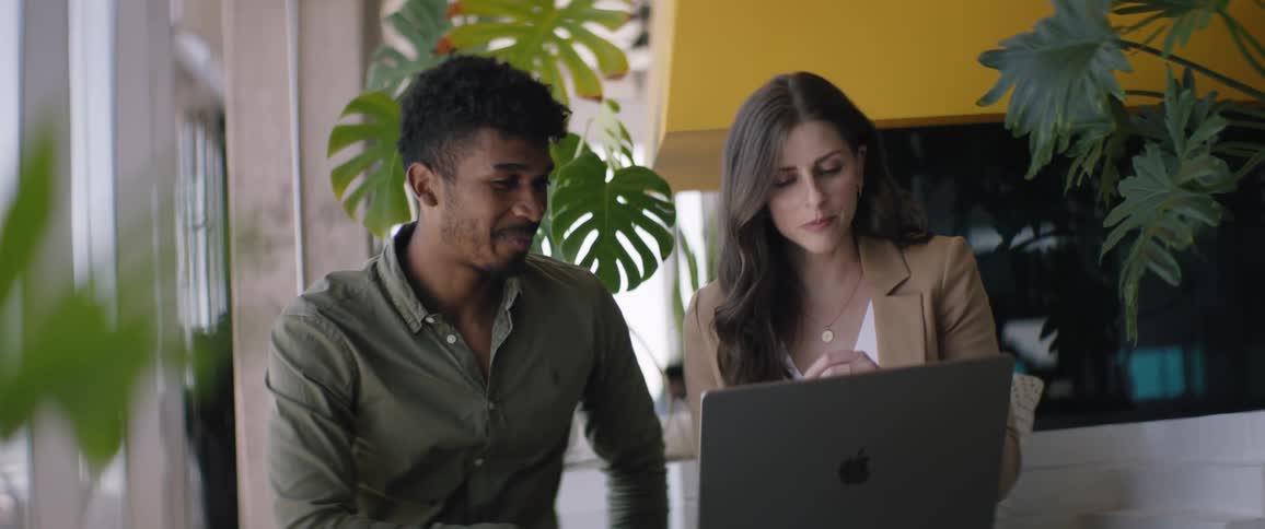 Woman and man sitting in a work environment talking and using a laptop