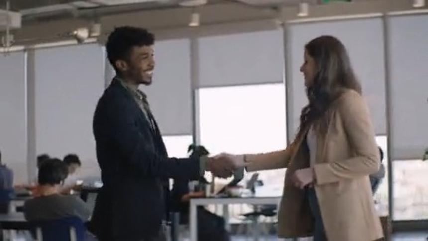 Man and woman greeting each other by holding hands and then sitting on an orange couch in an office