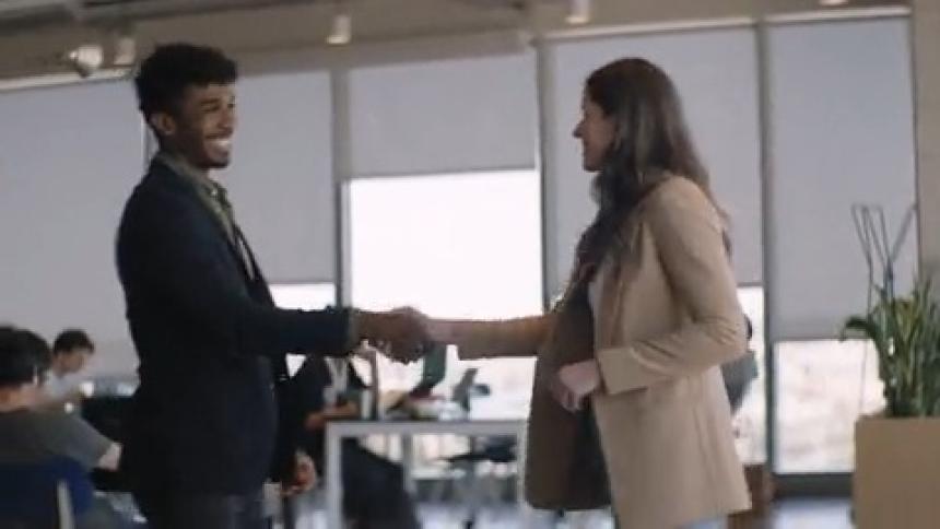 A man and a woman greet each other and then sit on a sofa and the woman starts talking while looking at a laptop