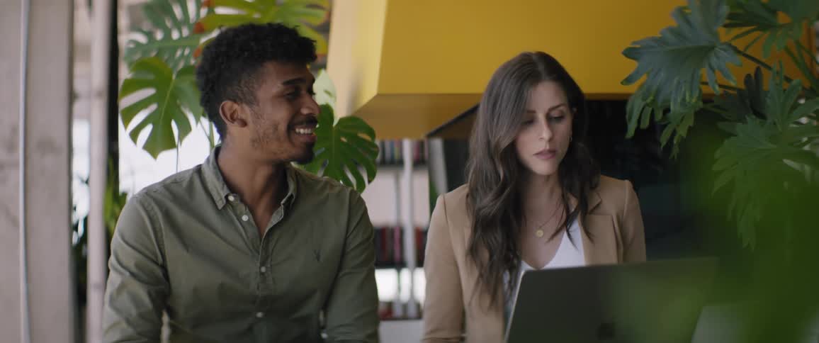 A man and a woman are sitting and talking while the woman is using the laptop in front of her