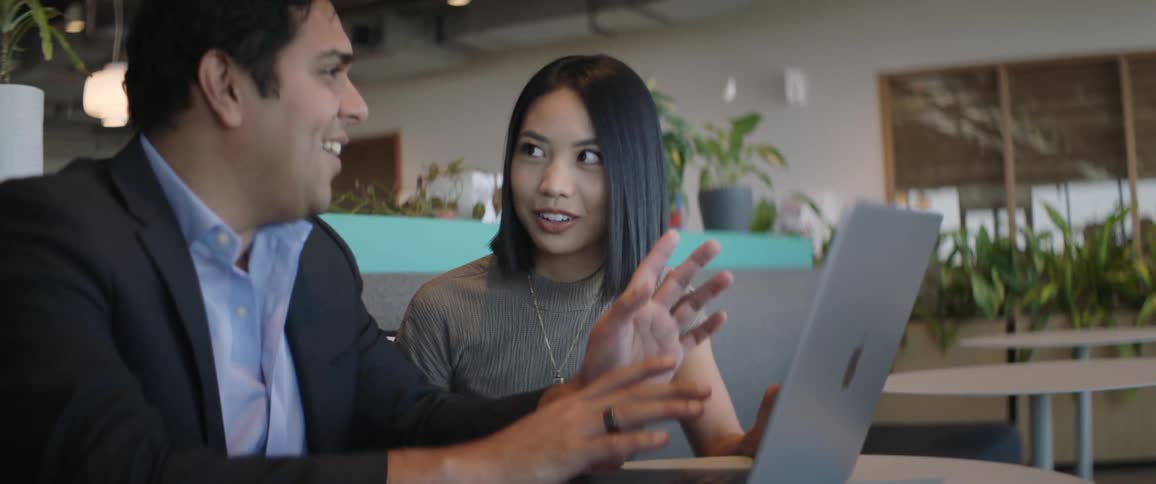 A man and a woman are sitting and talking while the man is typing on a laptop in front of them