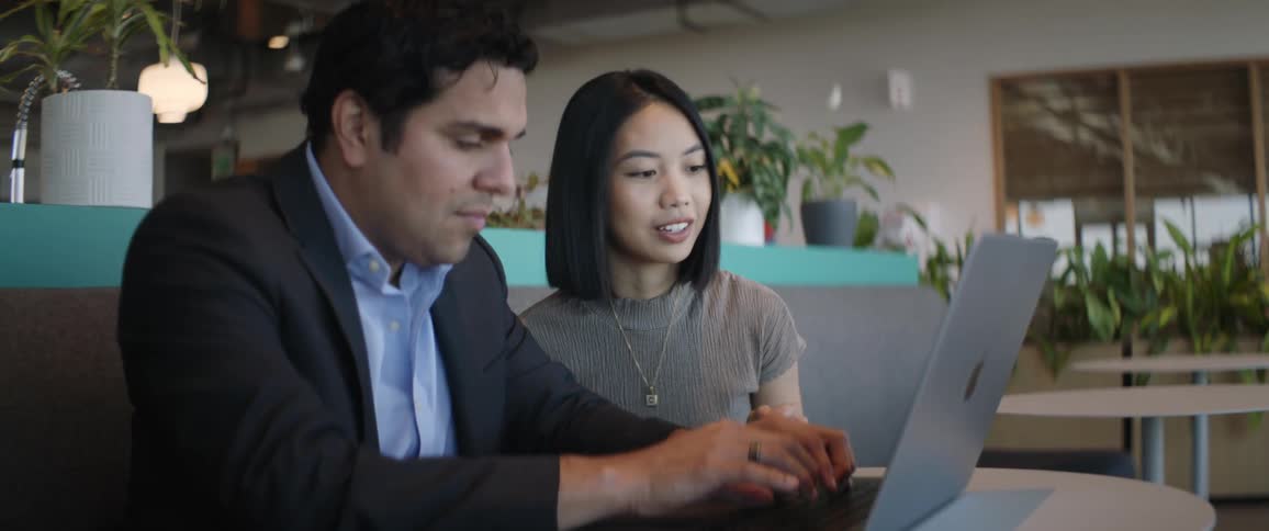 An asian woman speaks to an indian man as he types on a laptop