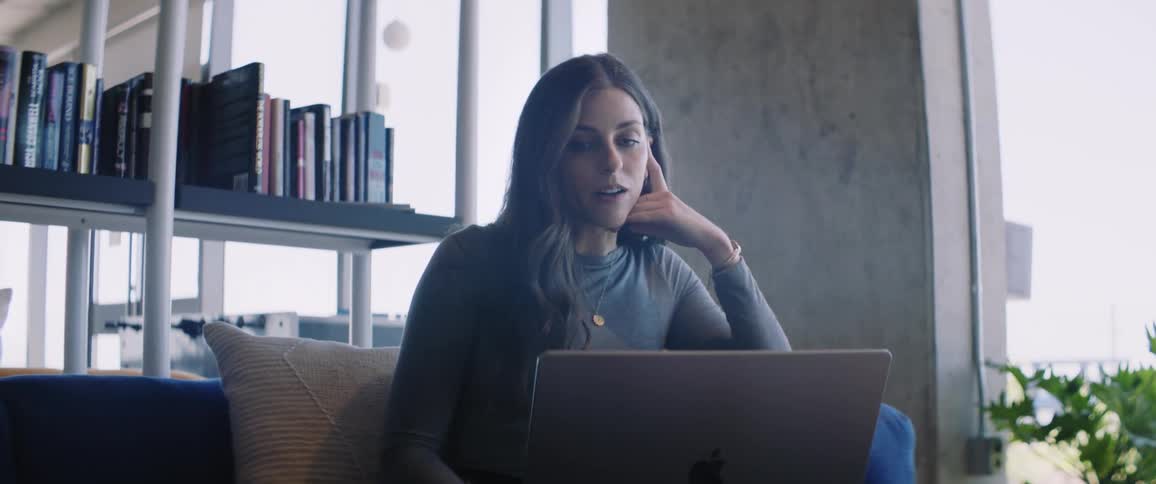 A young woman is sitting on the sofa and talking while looking at a laptop