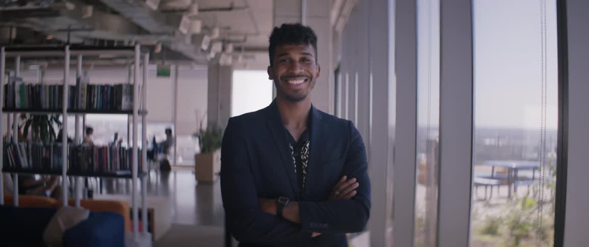 A black man wearing a jacket crosses his arms and smiles while standing in a library