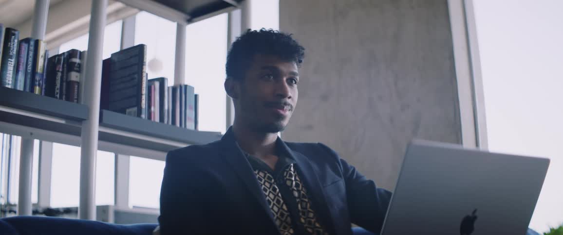 A young man wearing a jacket sits at his laptop computer near a shelf of books