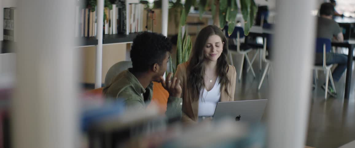 Man and woman sitting at a table in the library talk while working with a laptop