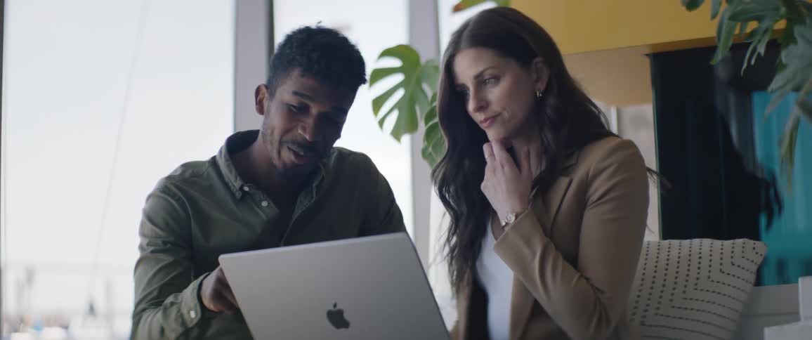 A man and a woman are talking while sitting and looking at a laptop that is on the womans legs