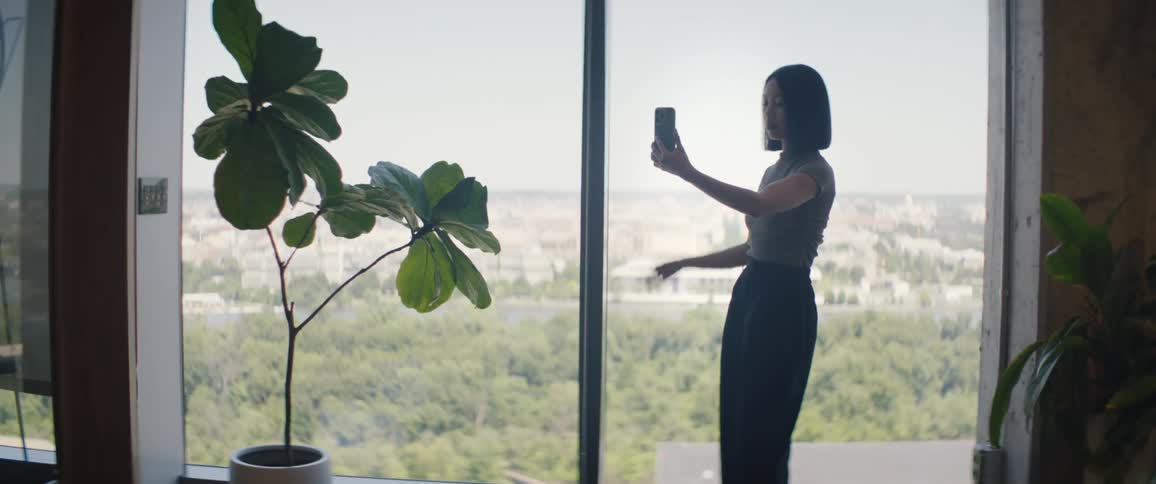 A young woman standing and talking with her cell phone pointed at her face next to a glass window
